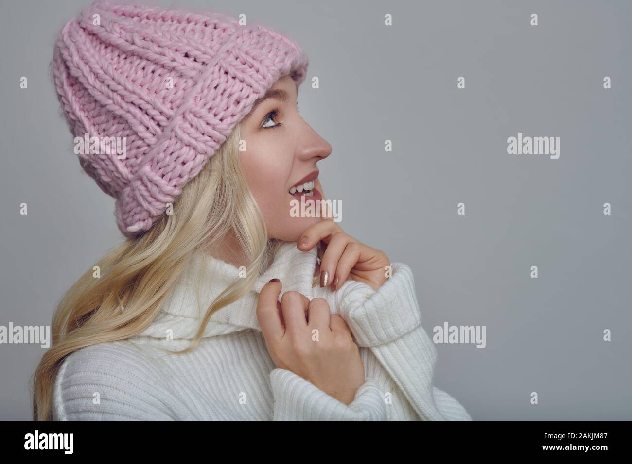 Pretty young blond woman in woollen winter outfit cuddling into the warmth of her polo neck sweater with a happy smile while wearing a pink knitted ca Stock Photo