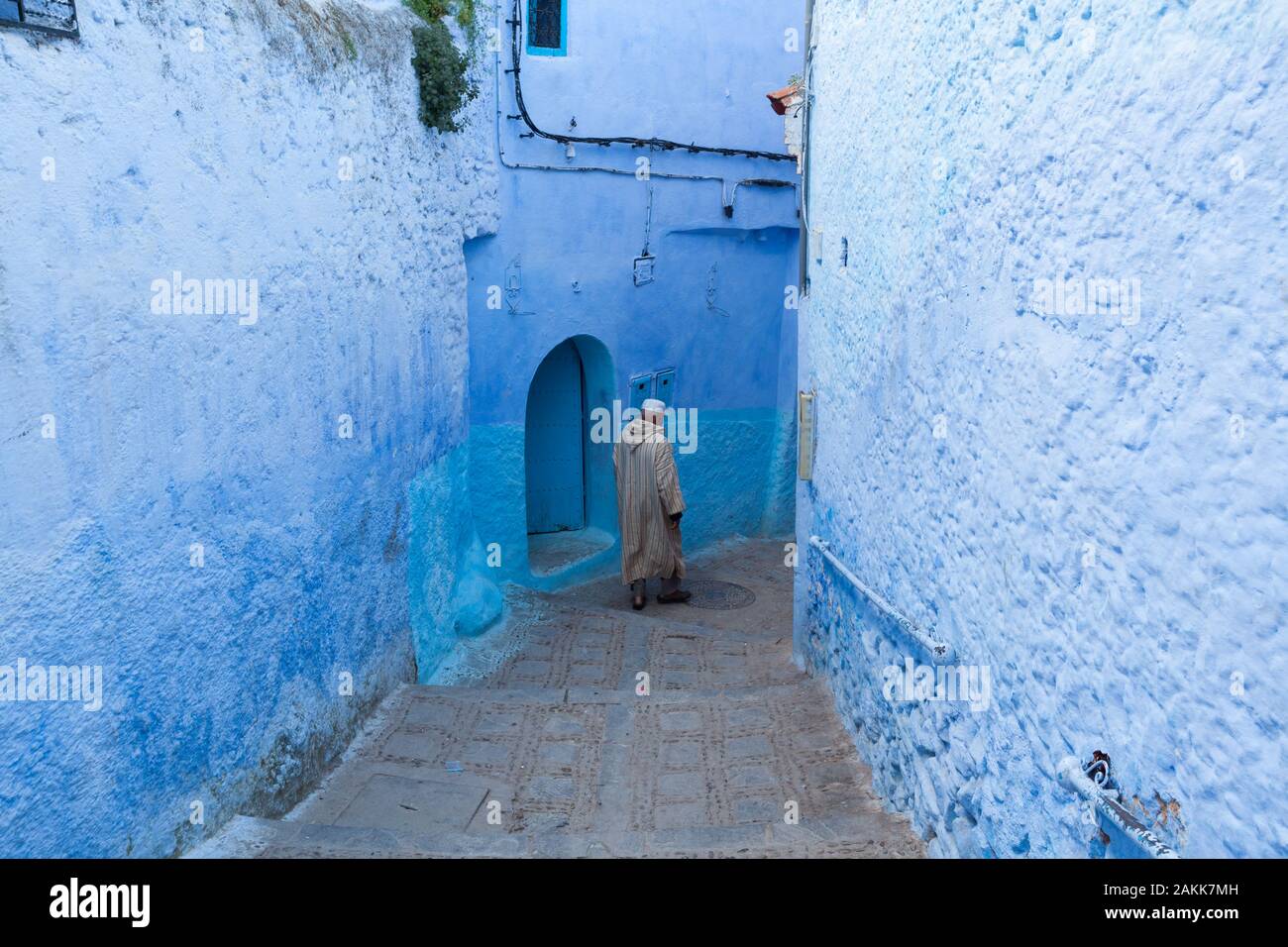 A solitary man in striped djellaba and white kufi cap on his head walking in the narrow street of medina in Chefchaouen, Morocco Stock Photo