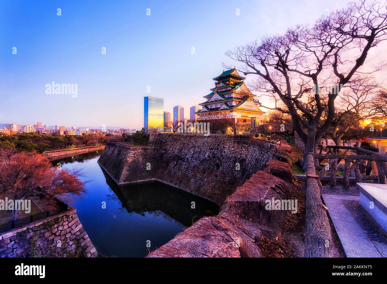 The main keep of Osaka castle from shogun times rising over stone walls and water filled moats at sunrise against clear sky and distant modern busines Stock Photo