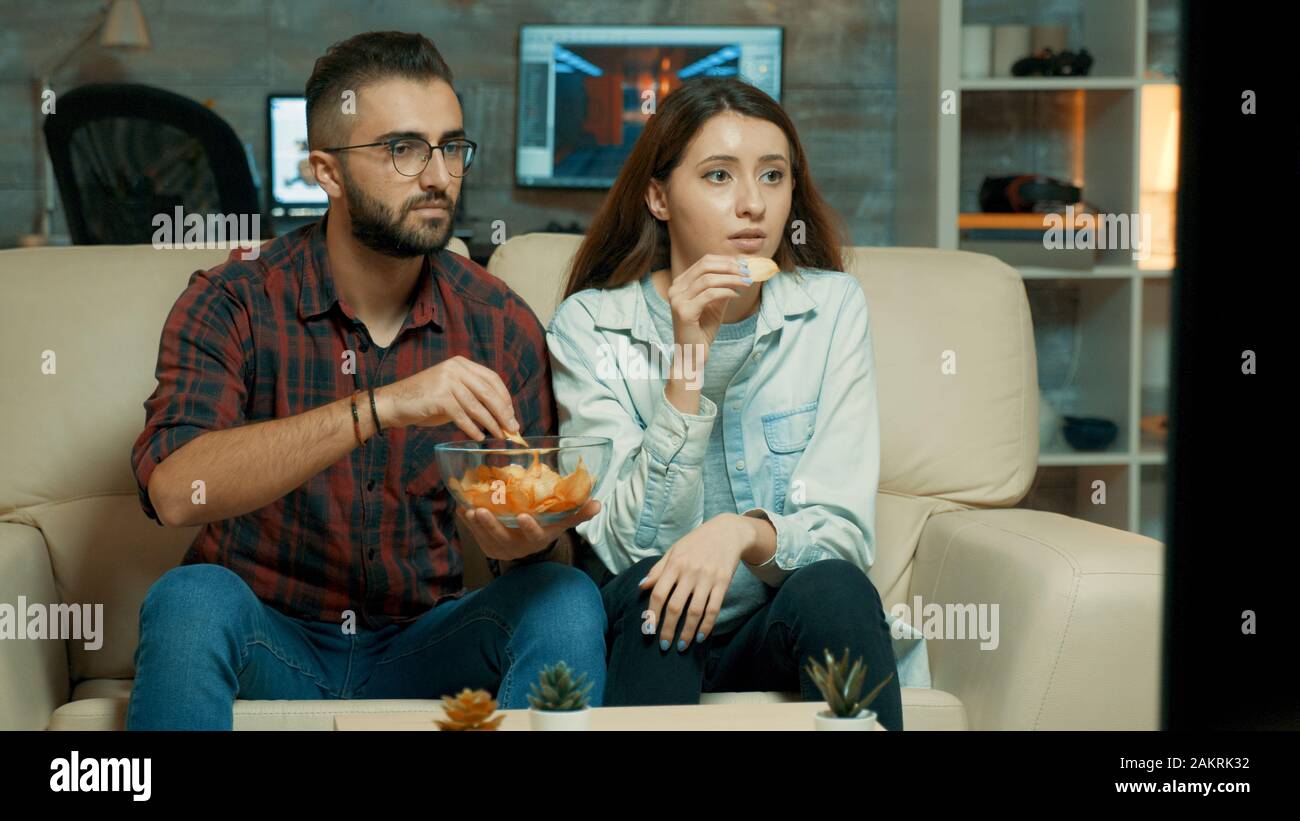 Concentrated caucasian young couple while watching tv and eating chips sitting on sofa. Stock Photo