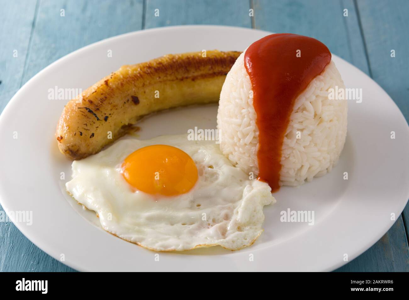 Arroz a la cubana Typical Cuban rice with fried banana and fried egg on a plate on wooden table. Stock Photo