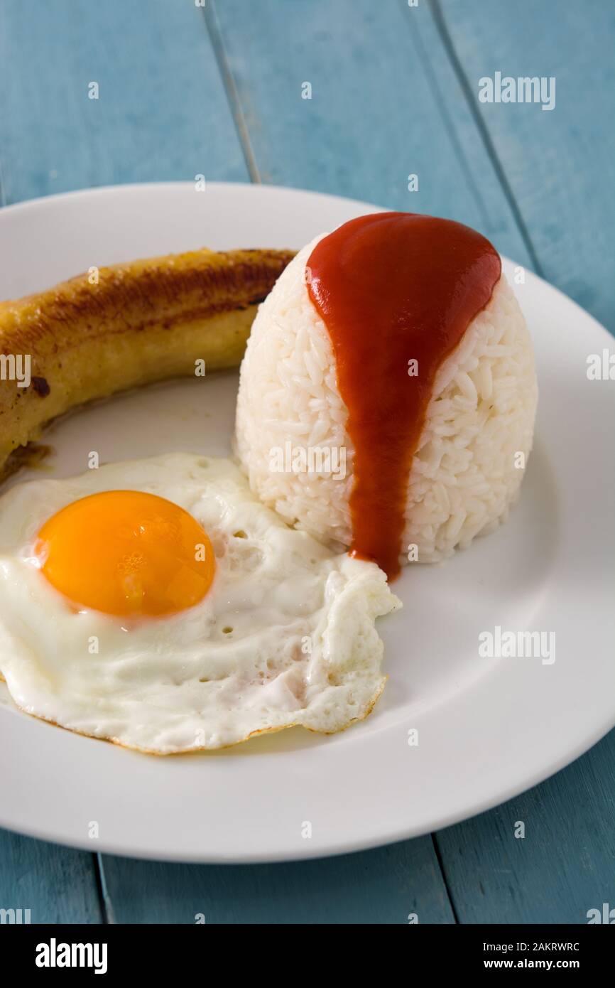 Arroz a la cubana Typical Cuban rice with fried banana and fried egg on a plate on wooden table. Stock Photo