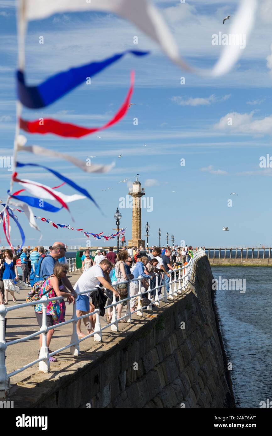 People on the Whitby West side harbour wall and lighthouse with royal red, white and blue bunting flying in the wind. Stock Photo