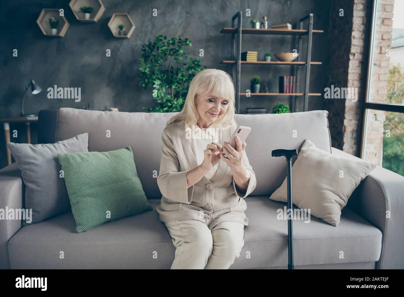 Portrait of her she nice attractive lovely cheery cheerful gray-haired grandma sitting on divan typing sms to relatives retirement at industrial brick Stock Photo