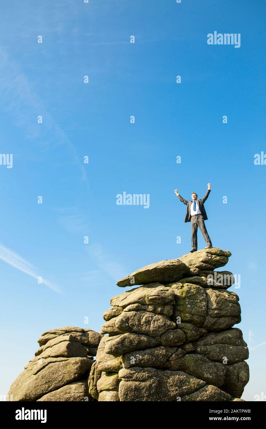 Businessman standing on the top of a distant rocky mountain peak celebrating in blue sky copy space Stock Photo