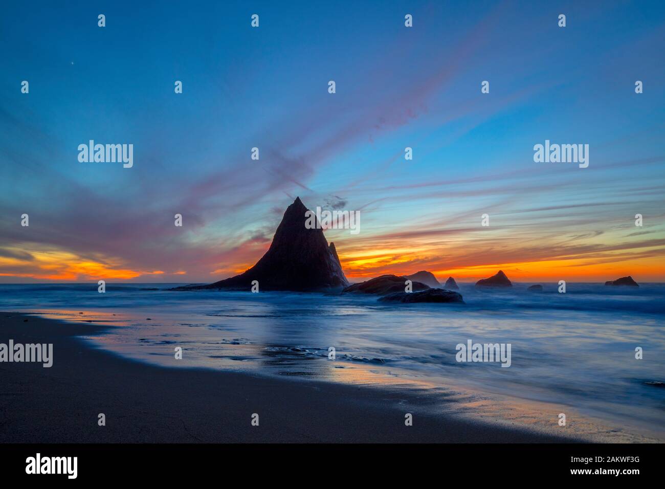 A colorful sunset backdrops the unique rock formations of Shark Fin Rock on Martin's Beach, in Half Moon Bay, California as the waves come in Stock Photo