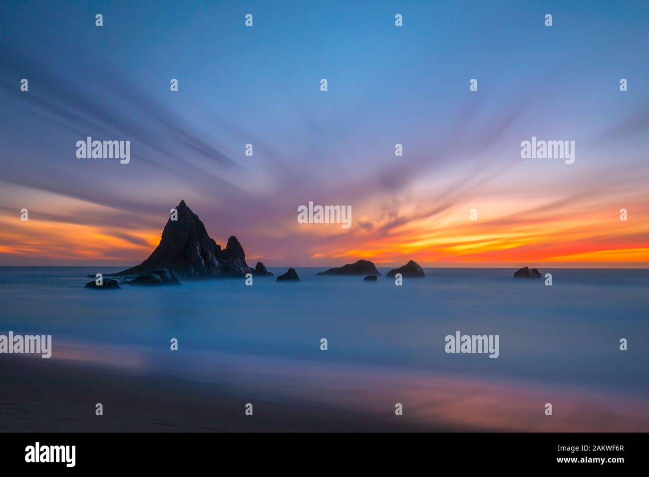 A colorful sunset backdrops the unique rock formations of Shark Fin Rock on Martin's Beach, in Half Moon Bay, California as the waves come in Stock Photo