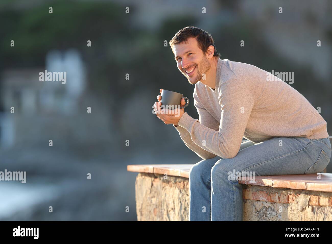 Happy man holding coffee cup contemplating views sitting in a balcony outdoors in a coast town Stock Photo