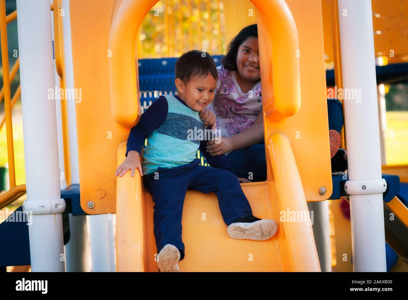 A little brother being helped by his older sister, who is about to go down a kids playground slide. Stock Photo