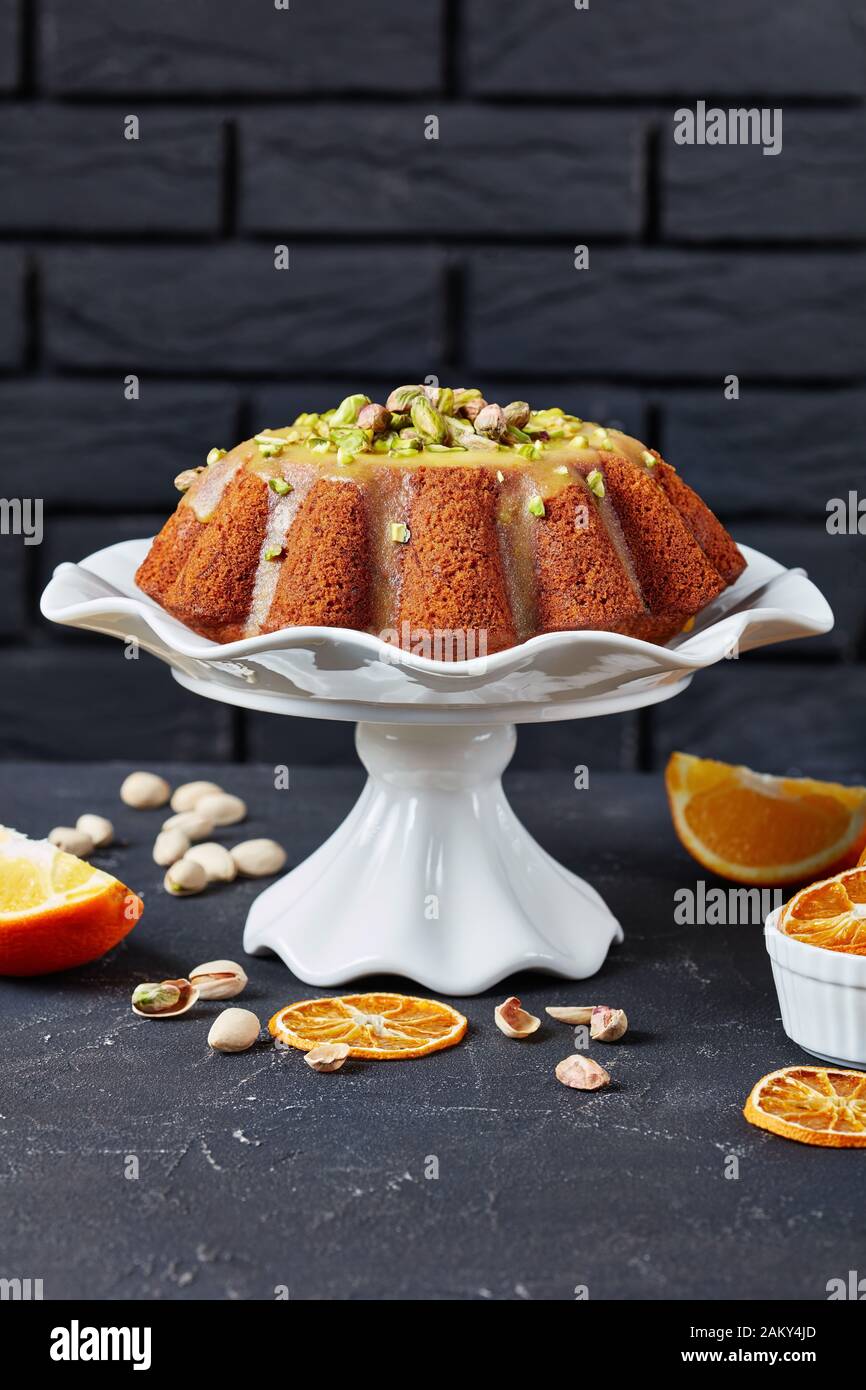 pumpkin carrot  bundt cake with orange glaze, sprinkled with pistachios on a white ceramic stand with a brick wall at the background and ingredients o Stock Photo
