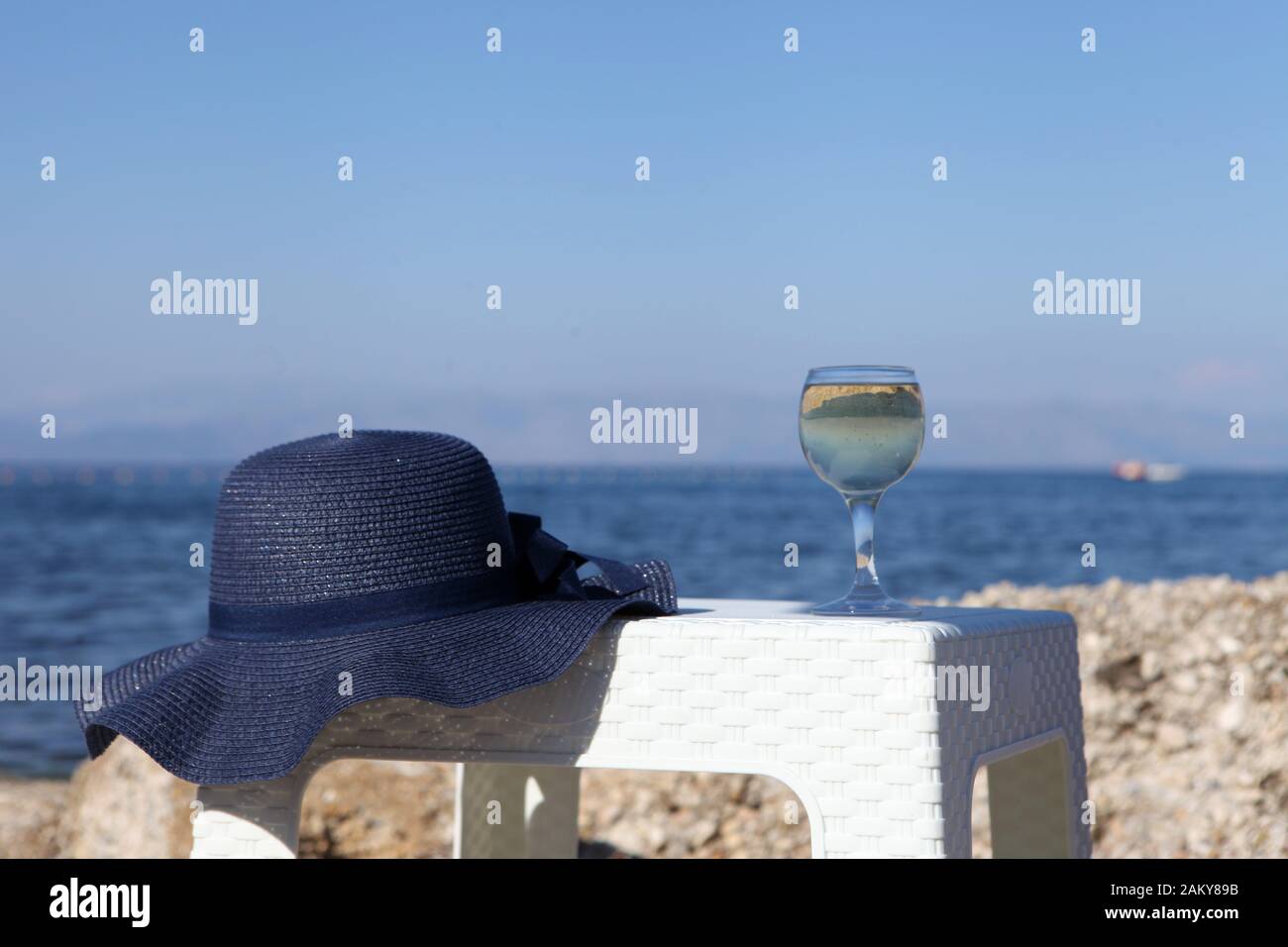 A glass of white wine and a blue hat behind lagoon sea. Summer vacation concept. Picnic with a glass of wine and blue hat near the sea Stock Photo