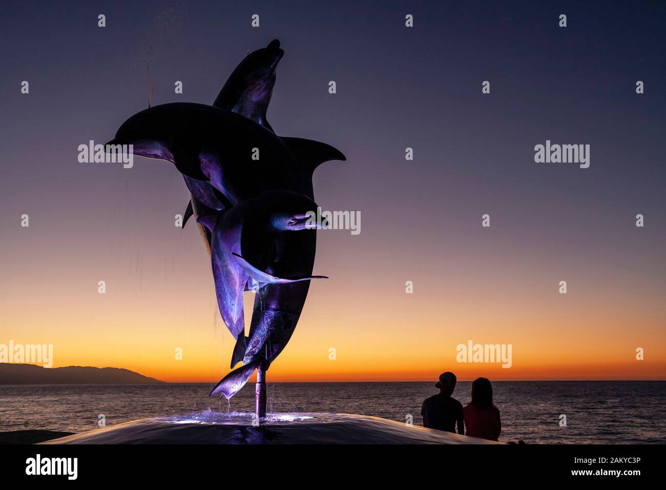 A Couple Sits and looks at the sunset over the ocean by the Friendship Fountain, the famous dolphin statue on the Malecon in Puerto Vallarta. Stock Photo