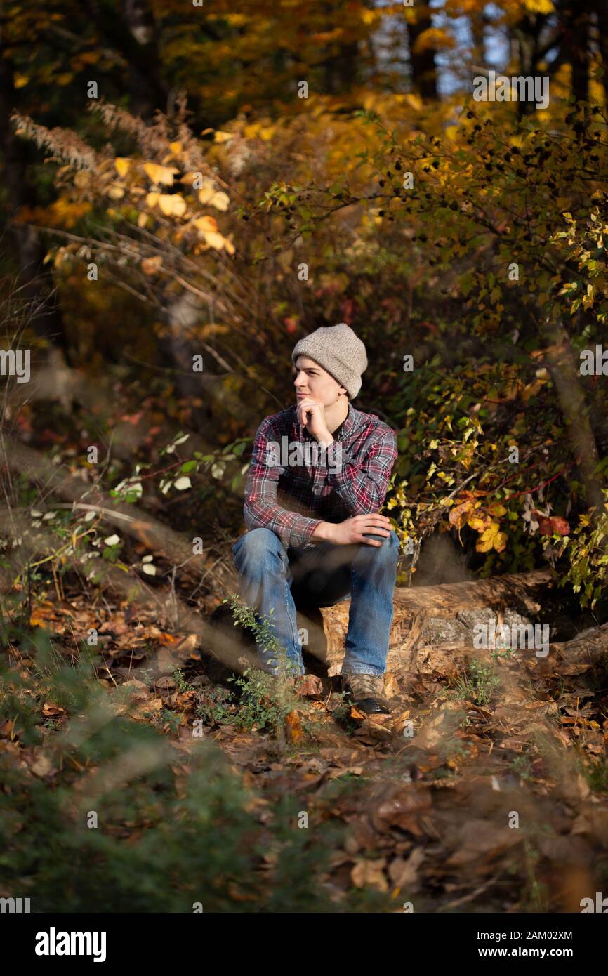 Teen boy sits alone on log in woods looking out thoughtfully. Stock Photo