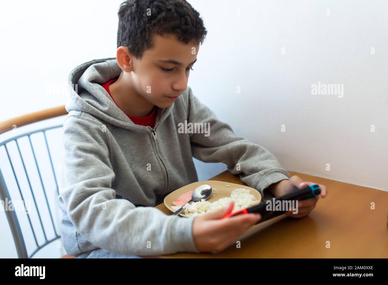boy eats poorly distracted by the game. Kid eating rice and surfing on internet or playing video games on console Stock Photo