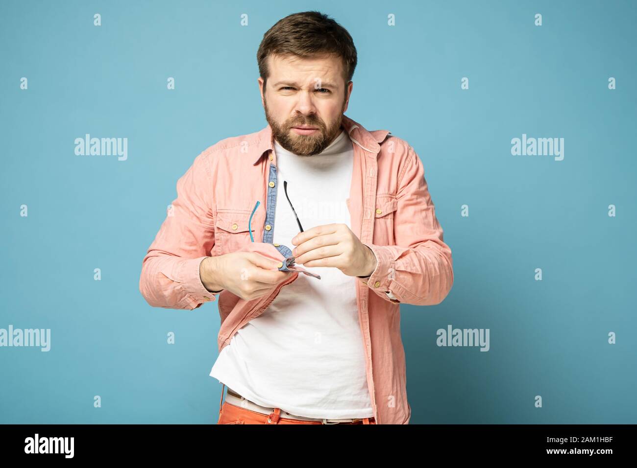 Bearded man cleans glasses, wiping them with a shirt and squinting, looking confidently into the camera. Stock Photo