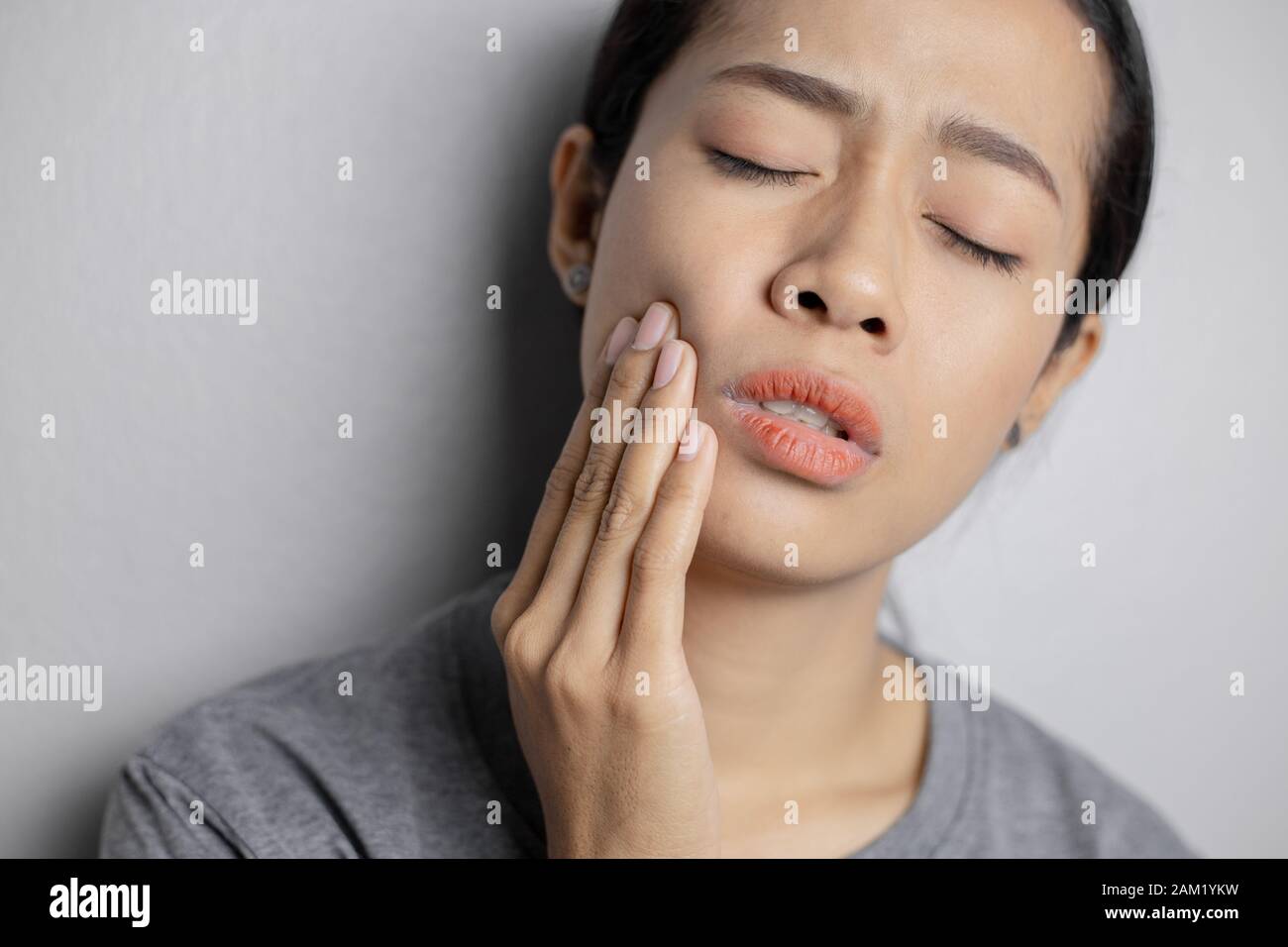 Woman put her hand on her cheek due to toothache. Asian  woman is suffering of toothache. Young woman with of toothache on a gray background. Stock Photo