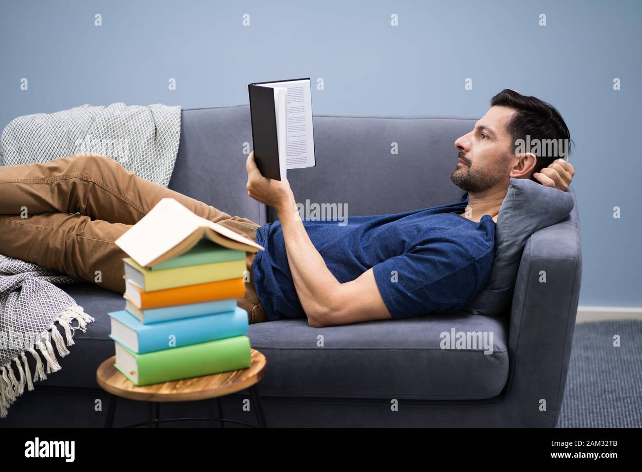 Man Lying On Sofa Reading Book At Home Stock Photo