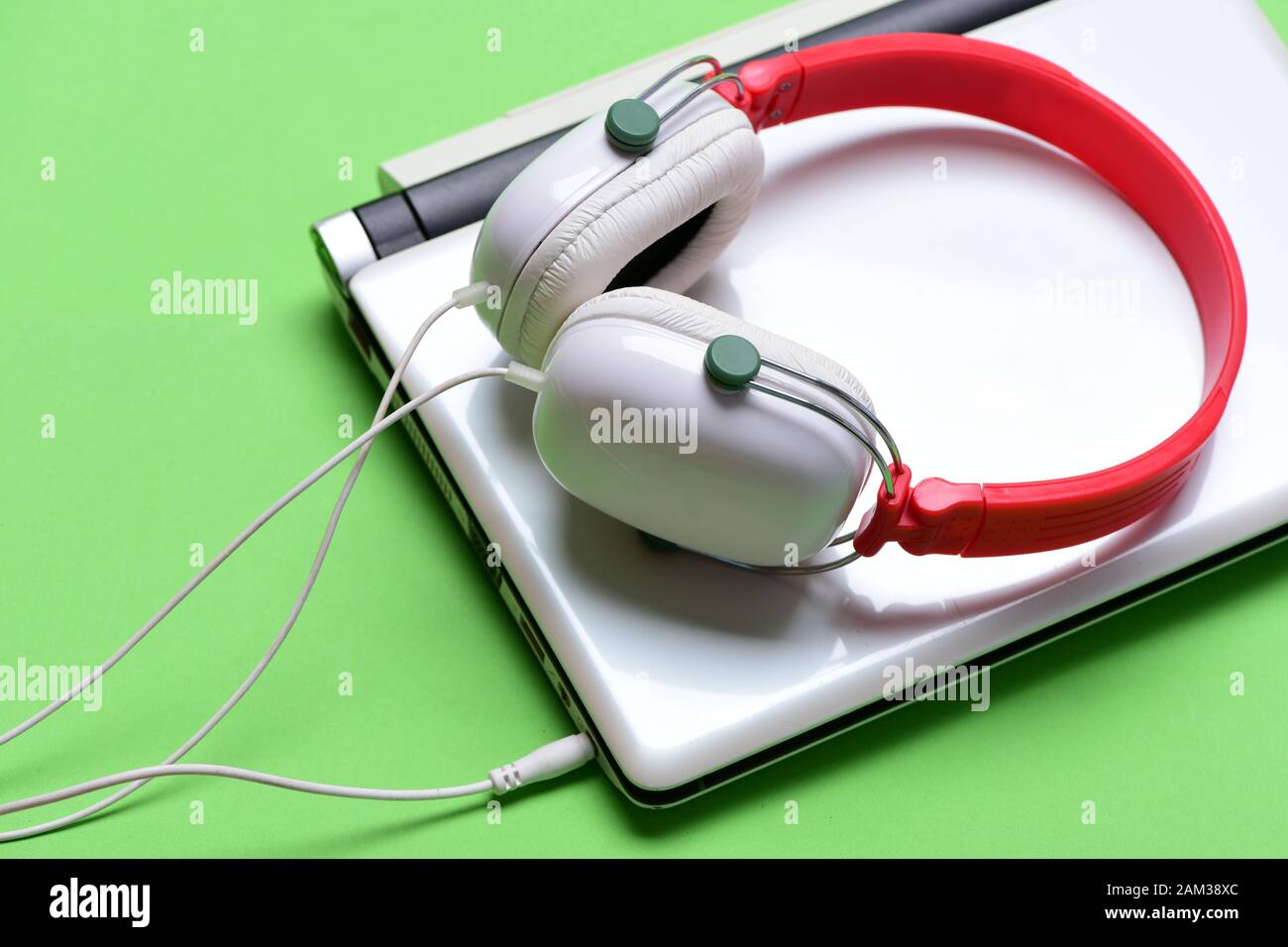 Headphones and silver laptop. Music and digital equipment concept. Electronics isolated on green background. Earphones in red and white colors made of plastic with computer. Sound recording idea. Stock Photo