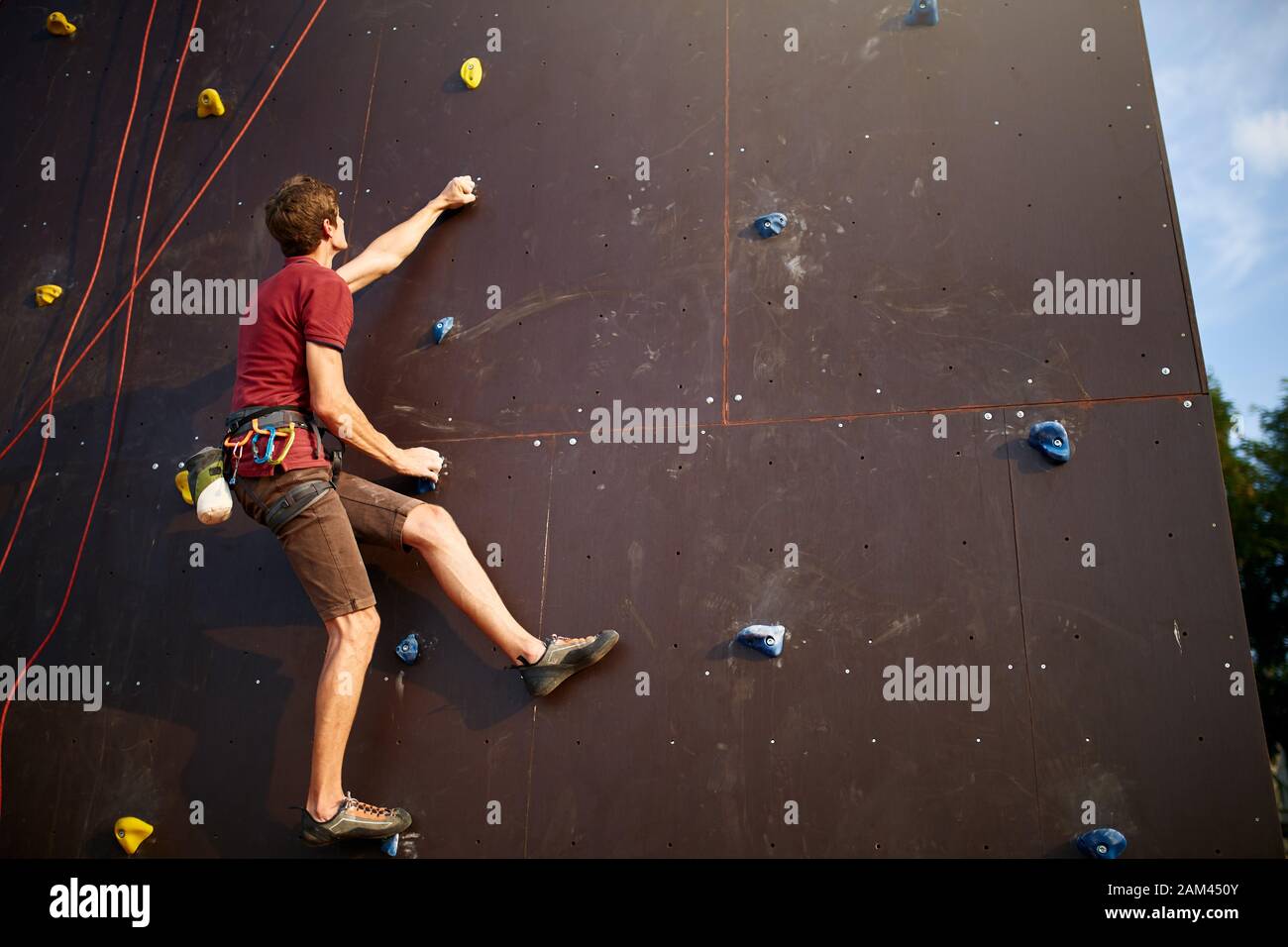 Sporty man practicing rock climbing in gym on artificial rock training wall outdoors. Young talanted slim climber guy on workout. Stock Photo