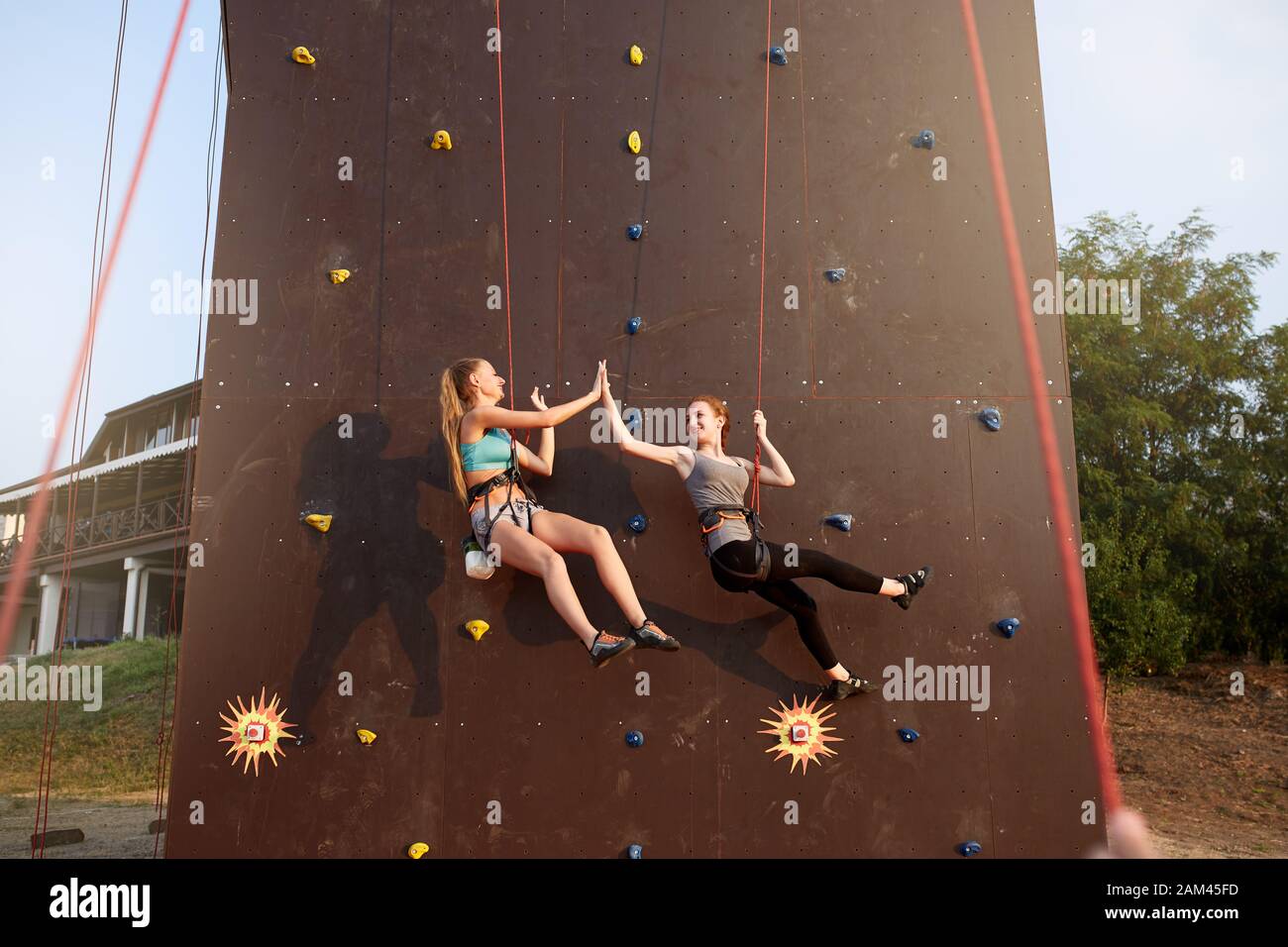 Smiling girls giving high five hanging on ropes at artificial climbing training wall and insured by friends on belaying harness. Rock climbing safety Stock Photo