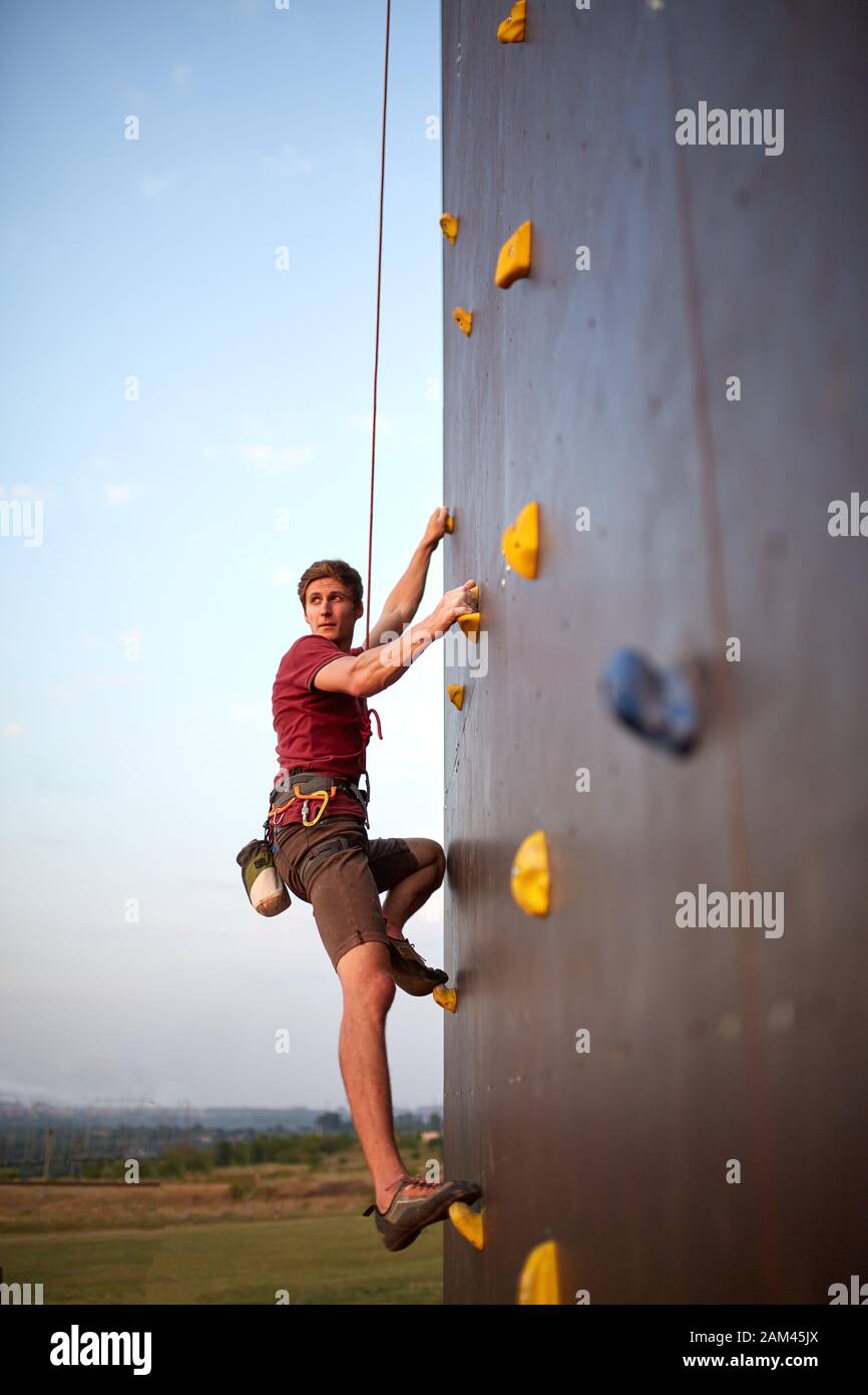 Sporty man practicing rock climbing in gym on artificial rock training wall outdoors. Young talanted climber guy on workout. Stock Photo