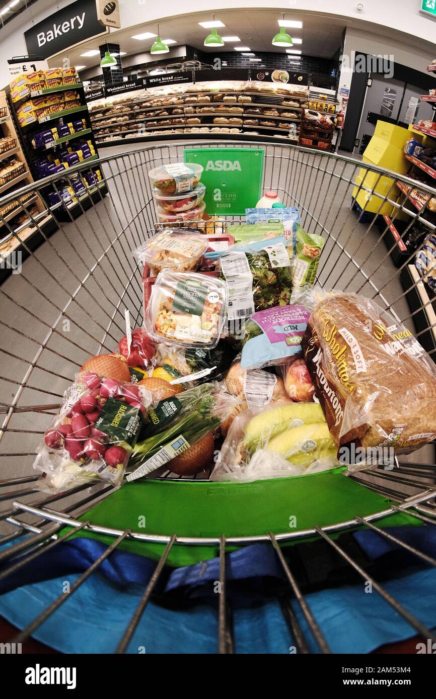 An Asda Supermarket grocery trolley or cart half full of plastic wrapped food items being pushed around a large supermarket. The Asda logo is visible Stock Photo