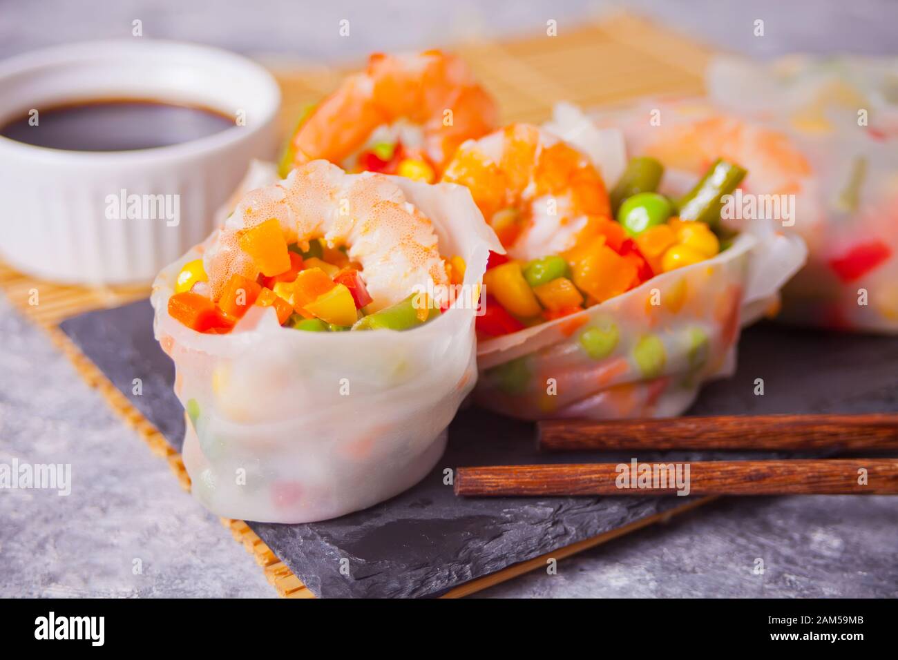 Spring rolls with vegetables and shrimps in a rice paper on the black plate. Near soy dip and chopsticks. Stock Photo