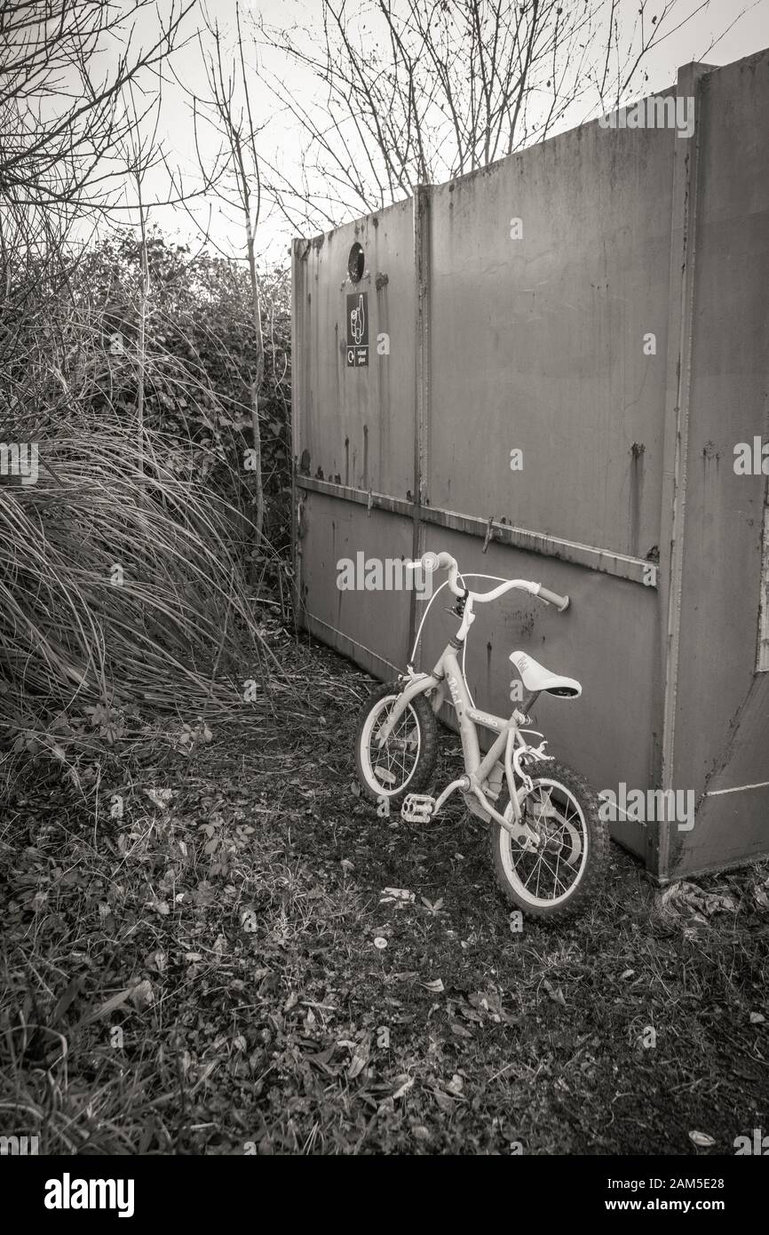 Balck and white of children's bike abandoned beside a recycling dumpster skip. Metaphor recycling, childhood memories, learning to ride a bike. Stock Photo