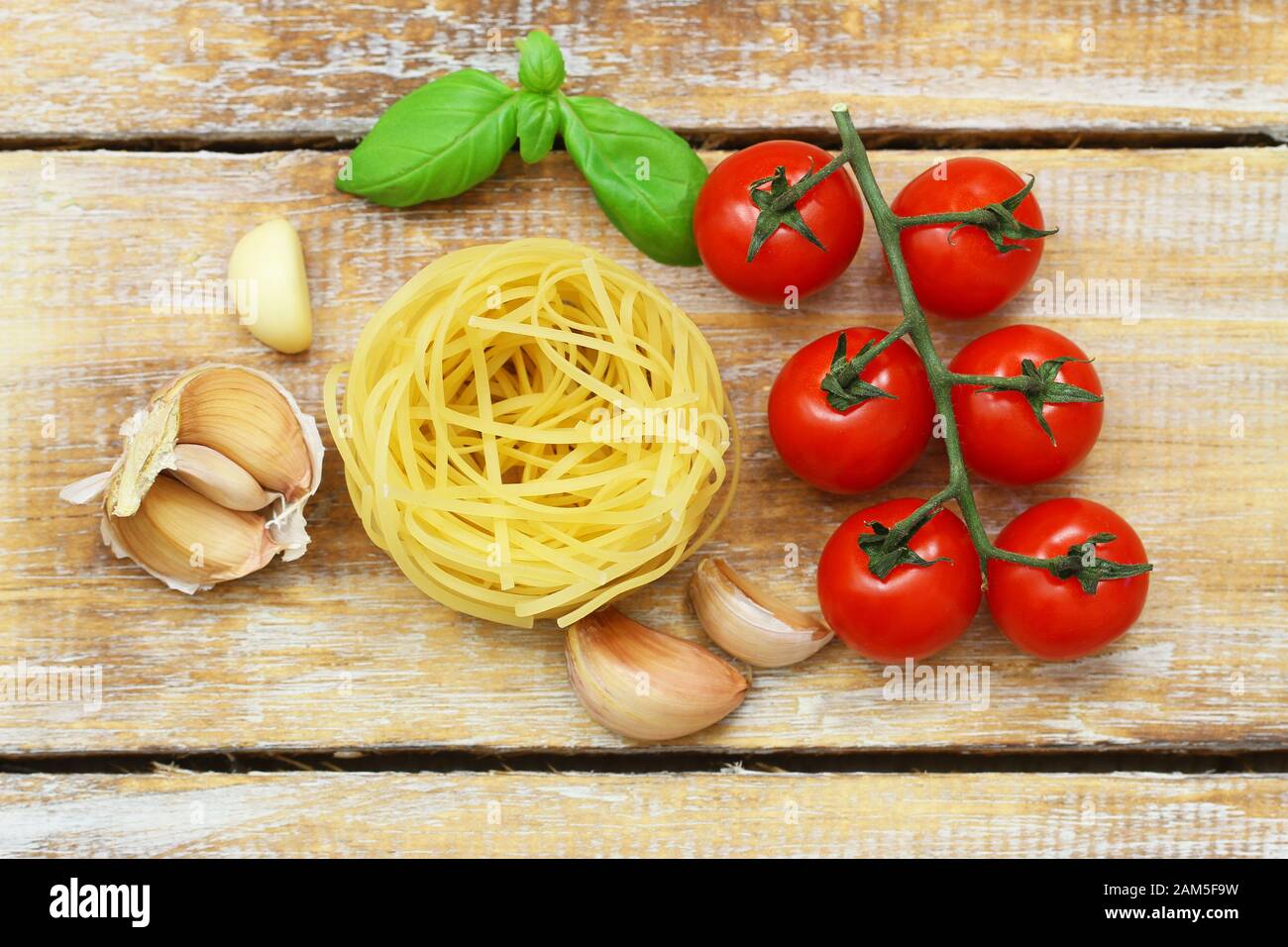 Ingredients for perfect Italian pasta: tagliatelle, ripe cherry tomatoes on stem, garlic cloves and fresh basil leaves on rustic wooden surface Stock Photo
