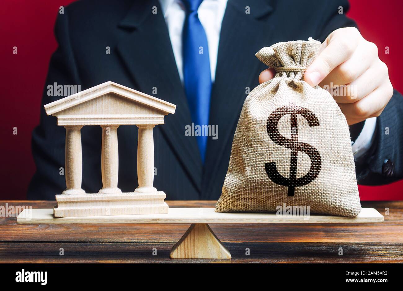 A man puts a money bag on scales opposite to building of government, bank, university. Budget and funding for normal functioning. Lobbying interests. Stock Photo