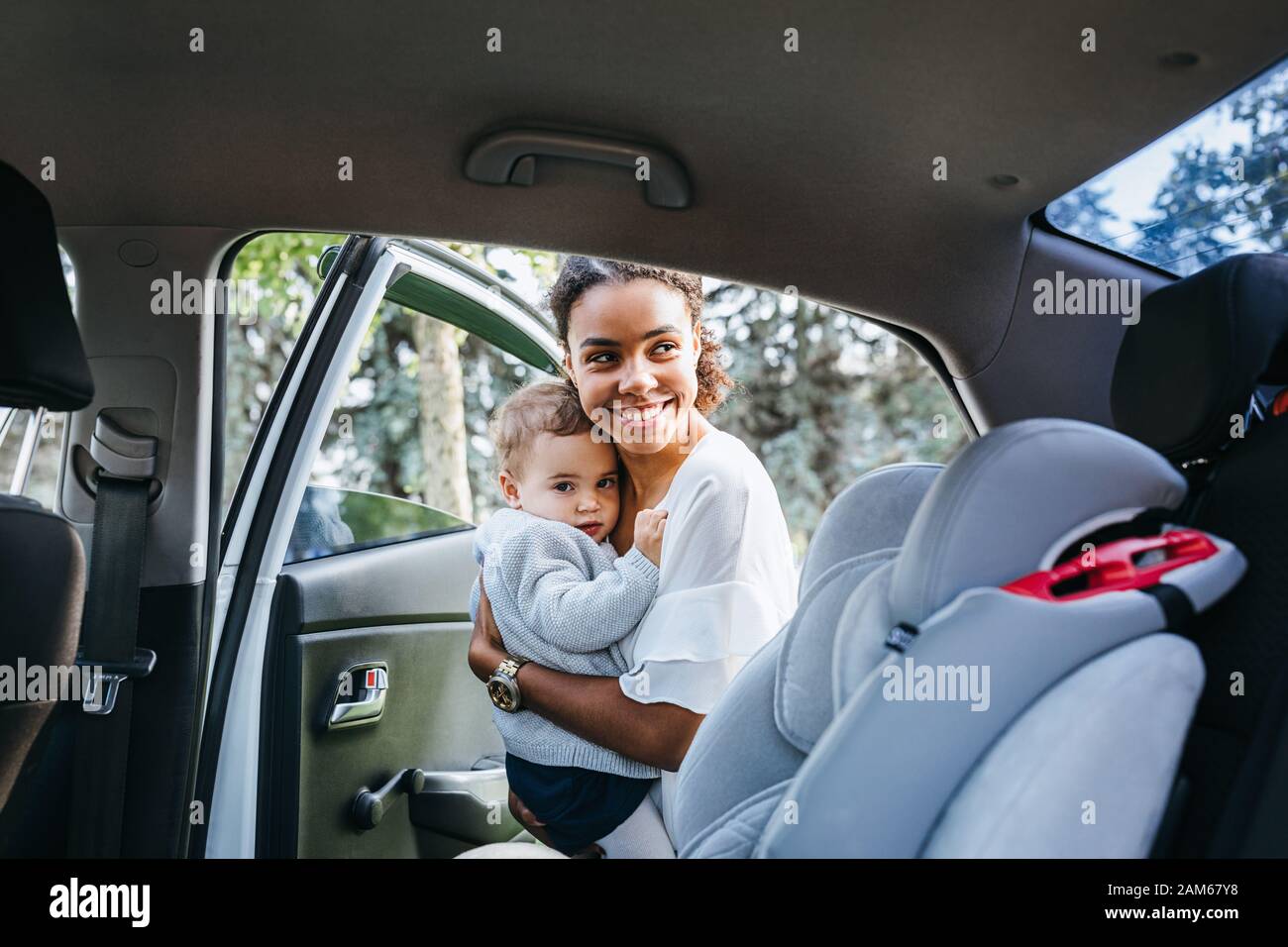 Smiling mother putting baby daughter in a car seat. Young woman holding a child on hands near a car. Stock Photo