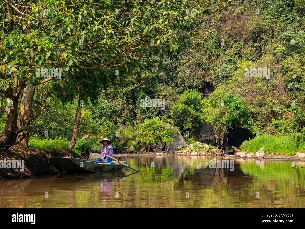 Local Asian woman wearing conical hat sitting in sampan on river with cavern entrance, Tam Coc cave system, Ninh Binh, Vietnam, Asia Stock Photo