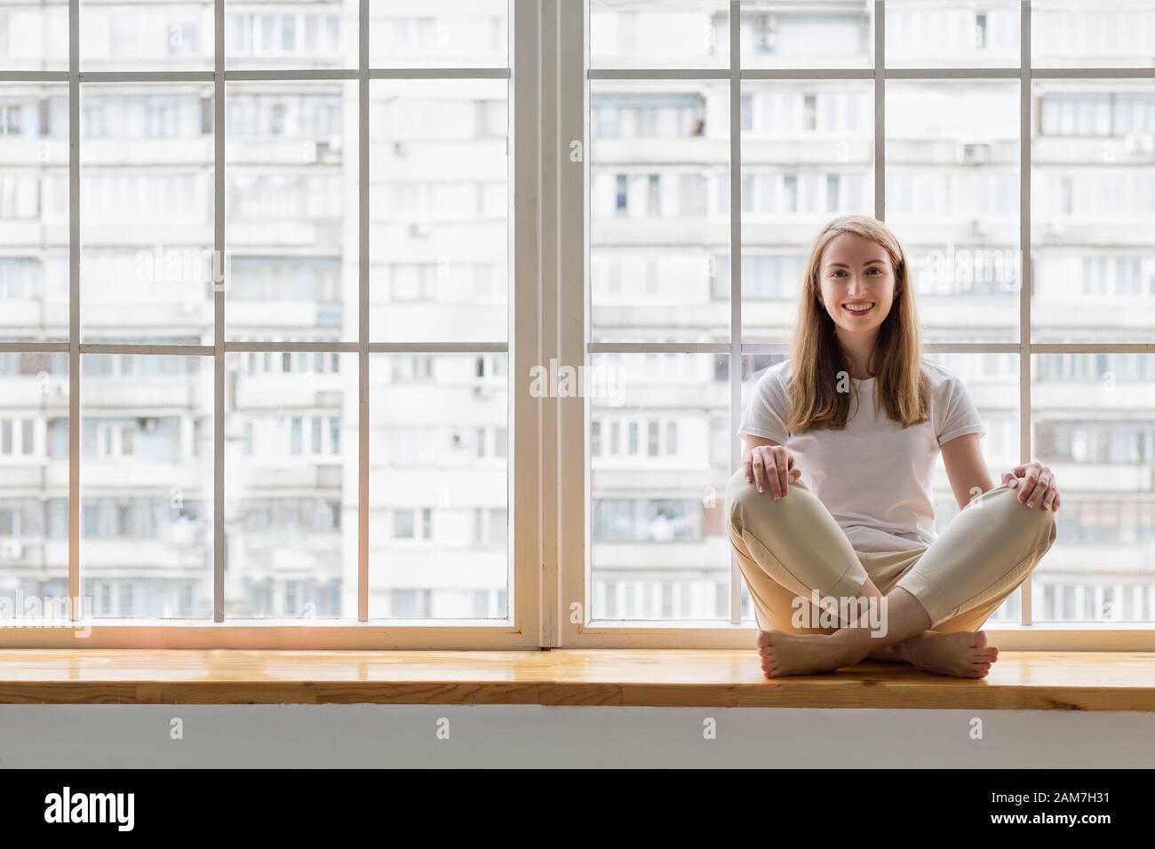 Happy young woman practicing yoga while sitting on window sill in lotus pose asana Stock Photo