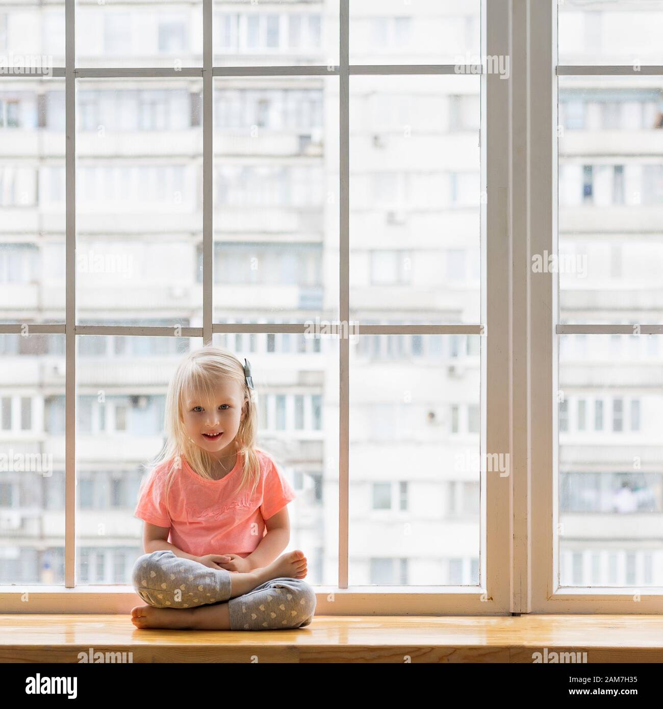 Baby girl doing yoga in front of window. Happy cute girl smiling while practicing yoga sitting in lotus pose asana Stock Photo