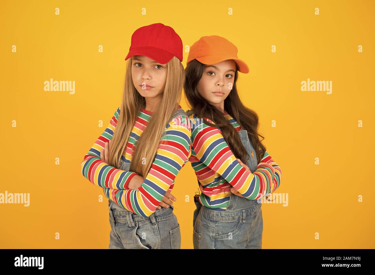 Fashion that matches their conscious style. Small girls in style keeping arms crossed on yellow background. Little children wearing baseball caps in casual streetwear style. Hip hop or hipster style S...