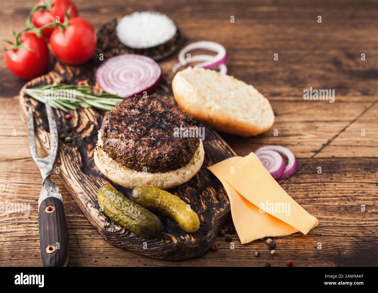 Fresh grilled minced pepper beef burger on vintage chopping board with buns onion and tomatoes on wood background. Salty pickles and fork Stock Photo