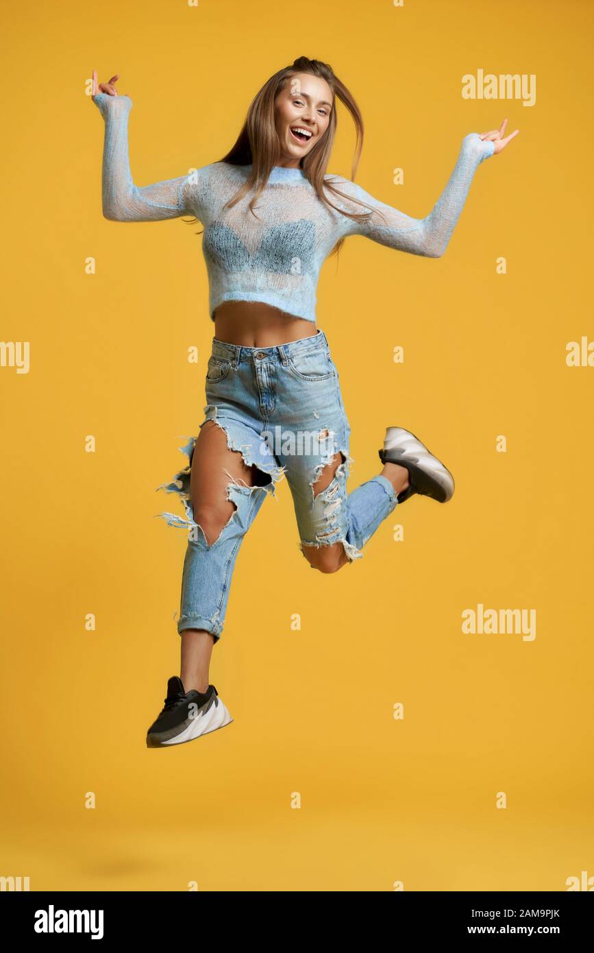 Front view of cheerful girl in full height with long brown hair raising  left leg up in jump wearing short blue top with long sleeves, ripped jeans  Stock Photo - Alamy