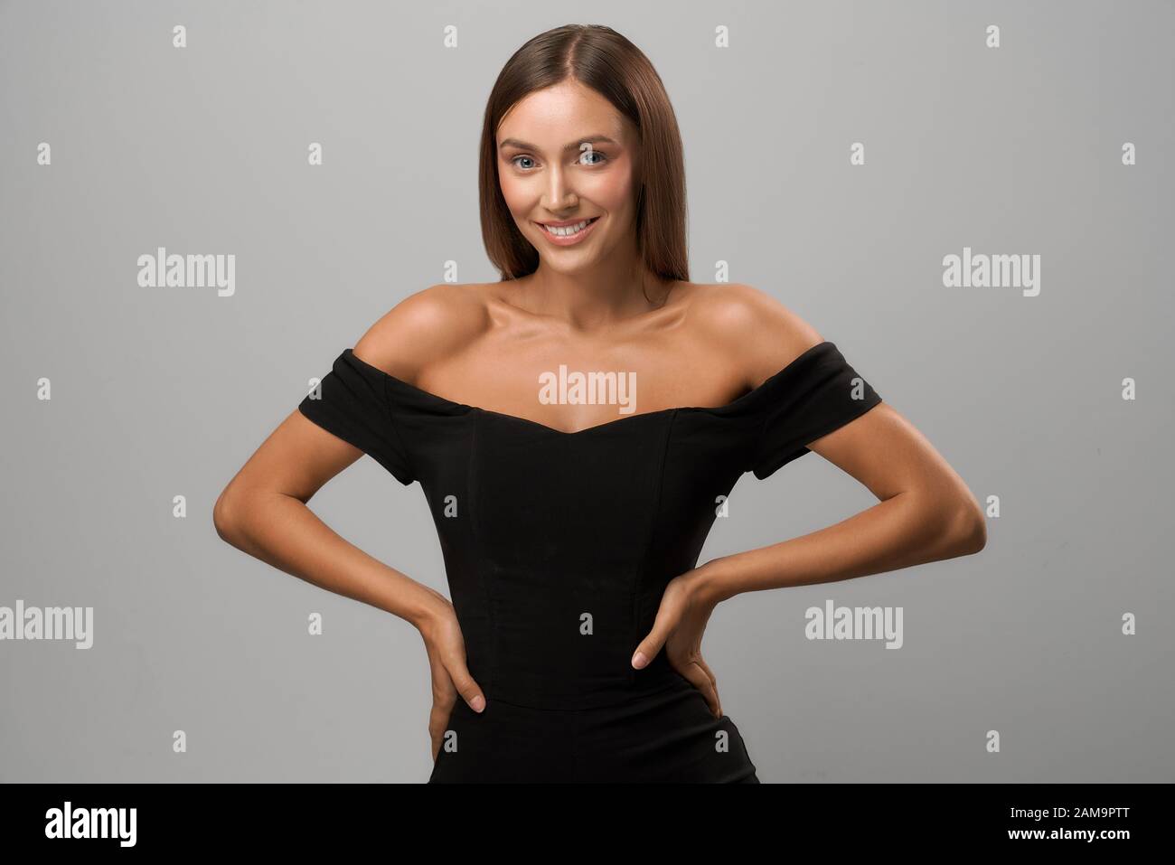 Front view of young attractive smiling girl with long brunette hair keeping hands on waist. Lady wearing black off shoulder top looking at camera on isolated grey background at studio. Stock Photo