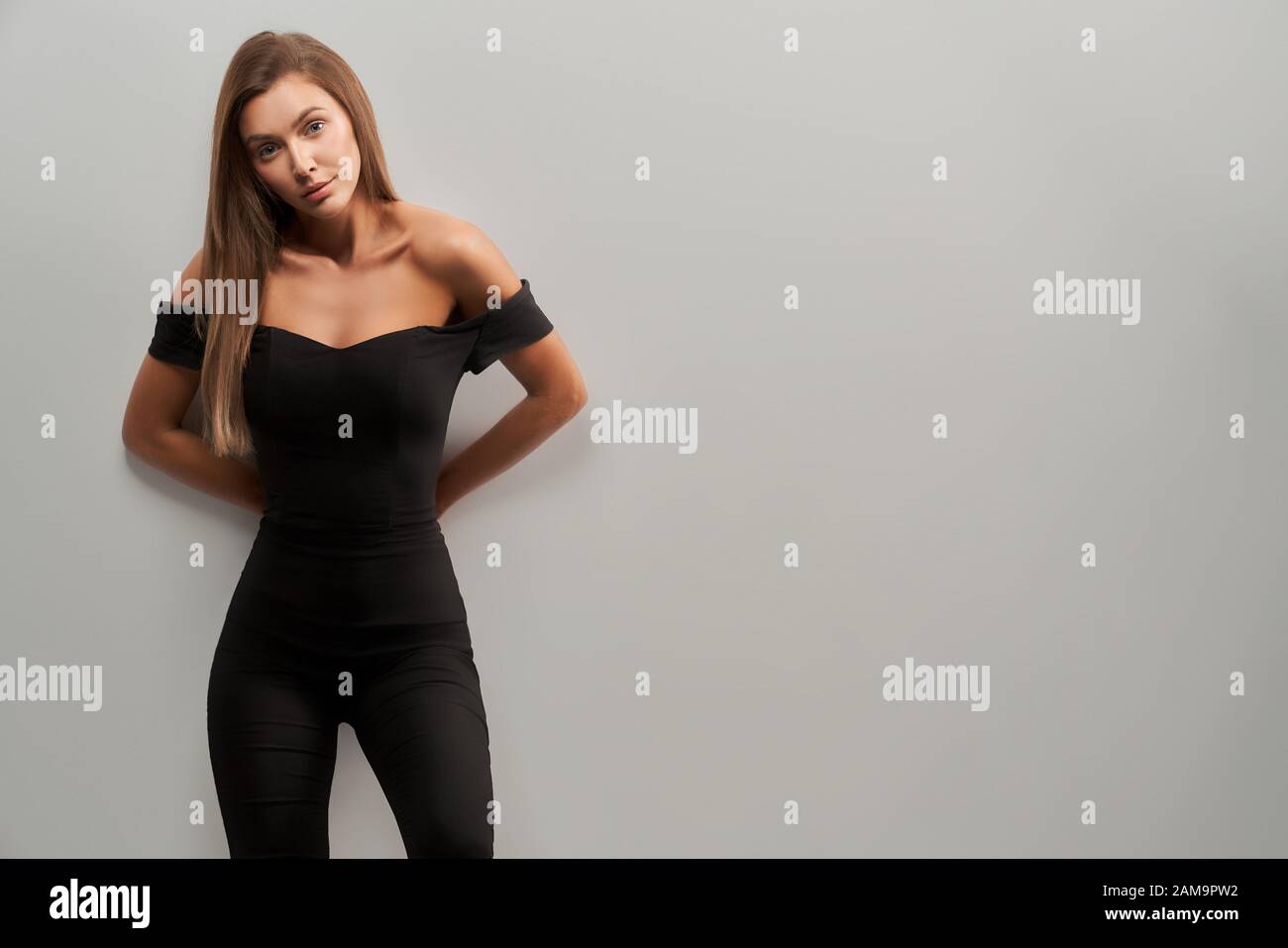 Front view of young attractive brunette girl with long hair posing tilting head and keeping hands behind back in black off shoulder top, trousers on isolated grey background at studio. Stock Photo