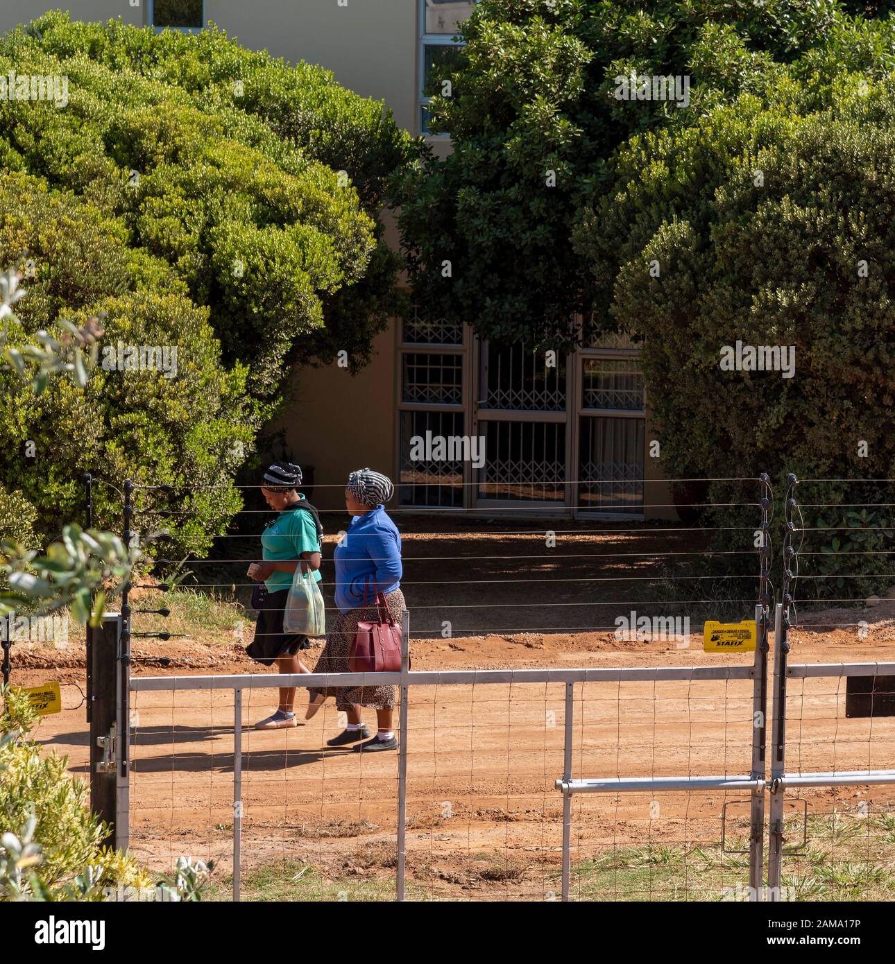 Western Cape, South Africa, December 2019. Two black women walking to work on a dirt road beyound a security fence. Stock Photo