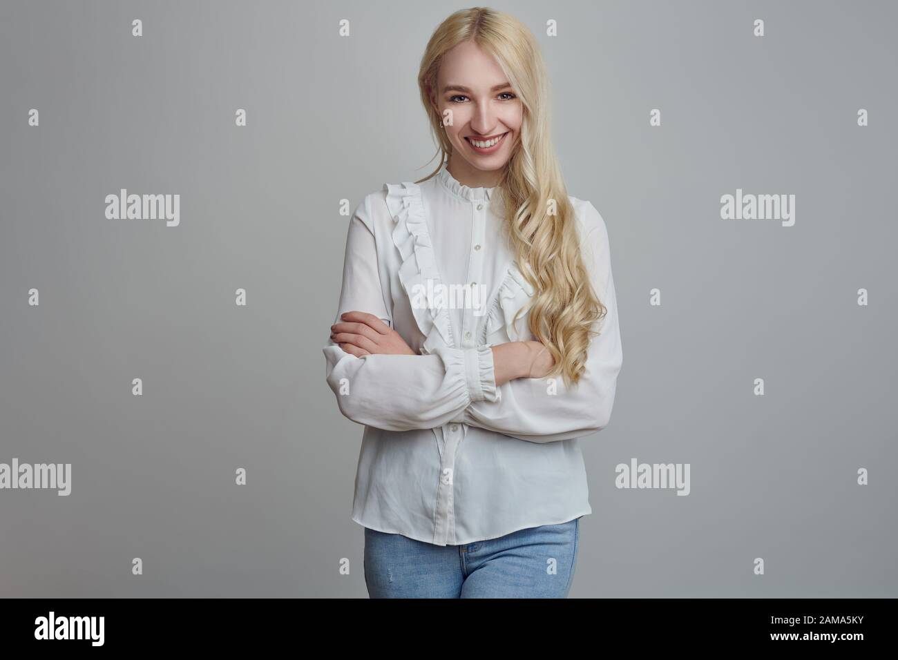 Young and beautiful long-haired blond woman in white blouse standing with her arms folded and looking at camera with a smirk. Front half-length portra Stock Photo