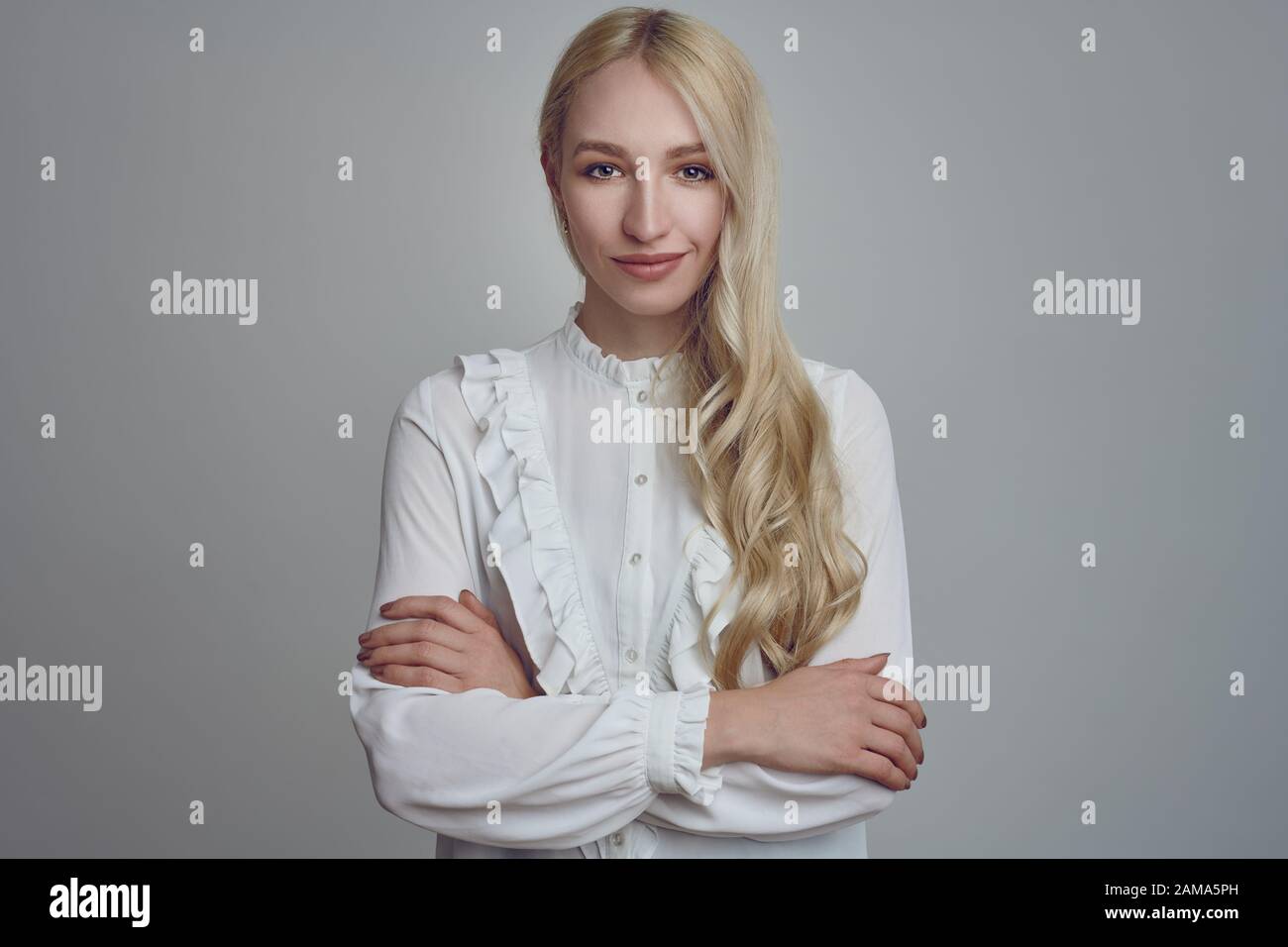 Young and beautiful long-haired blond woman in white blouse standing with her arms folded and looking at camera with a smirk. Front half-length portra Stock Photo