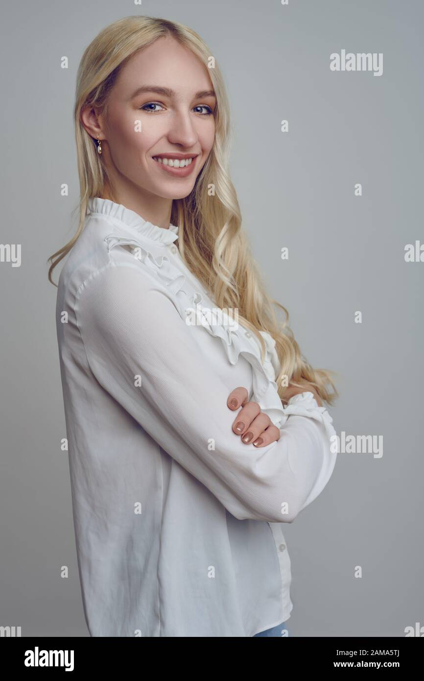 Young and beautiful long-haired blond woman in white blouse standing with her arms folded and looking at camera with a smile. Stock Photo
