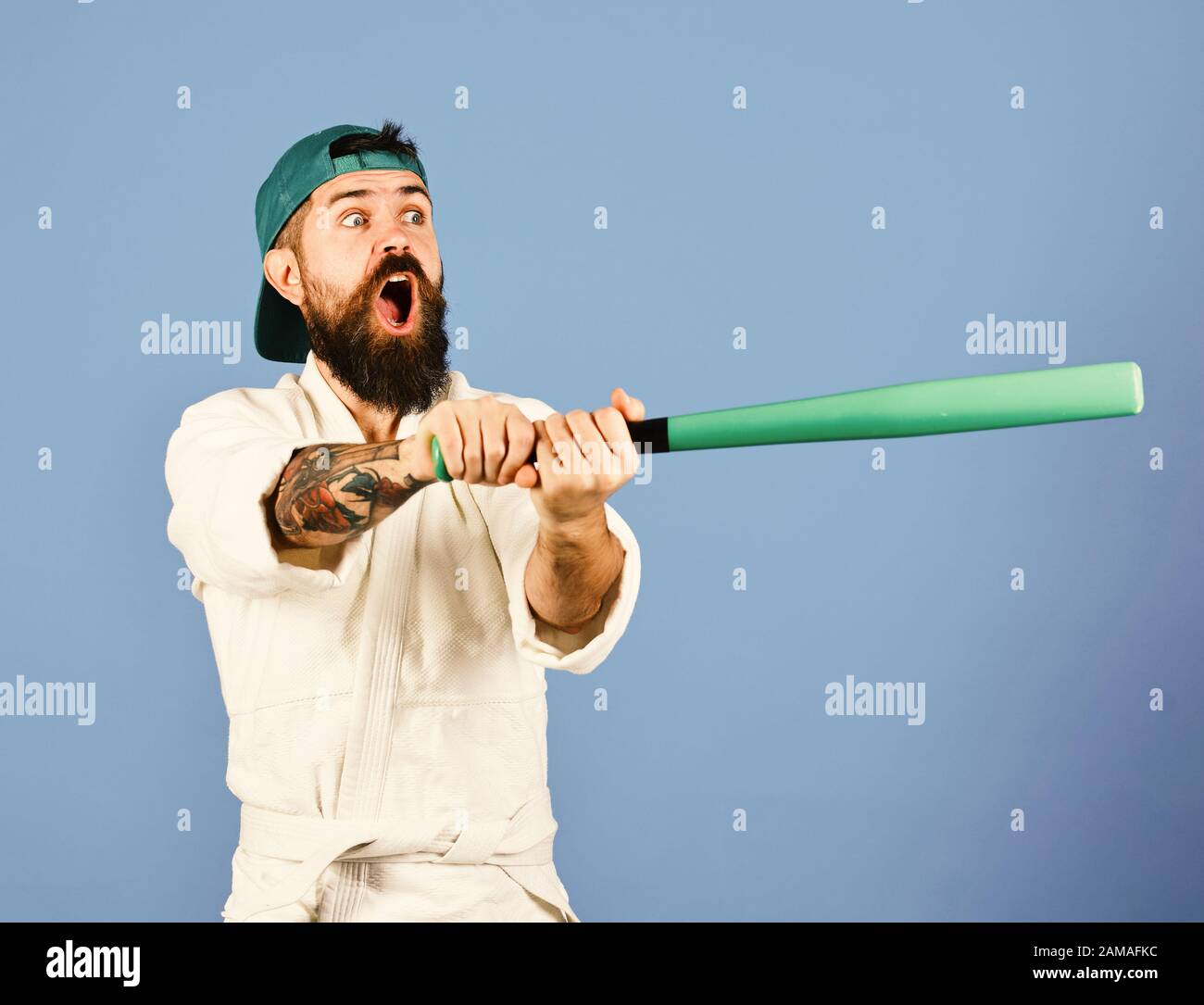 Man with beard in white and green on blue background. Martial arts Baseball player with shocked face holds green baseball bat. Master gets ready to fight Stock Photo -