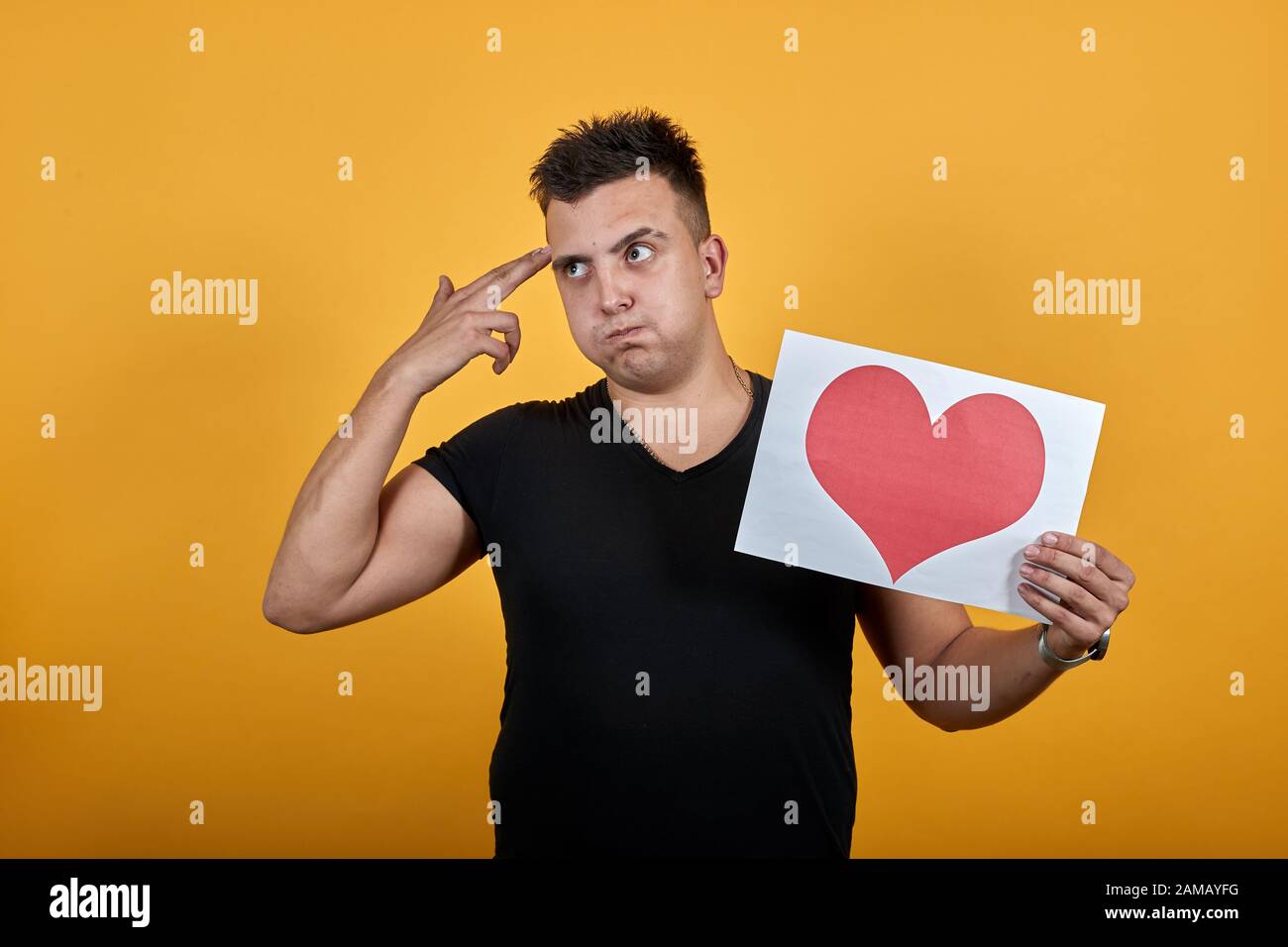 Unhappy young man keeping picture of heart, holding gun with fingers near head. Stock Photo