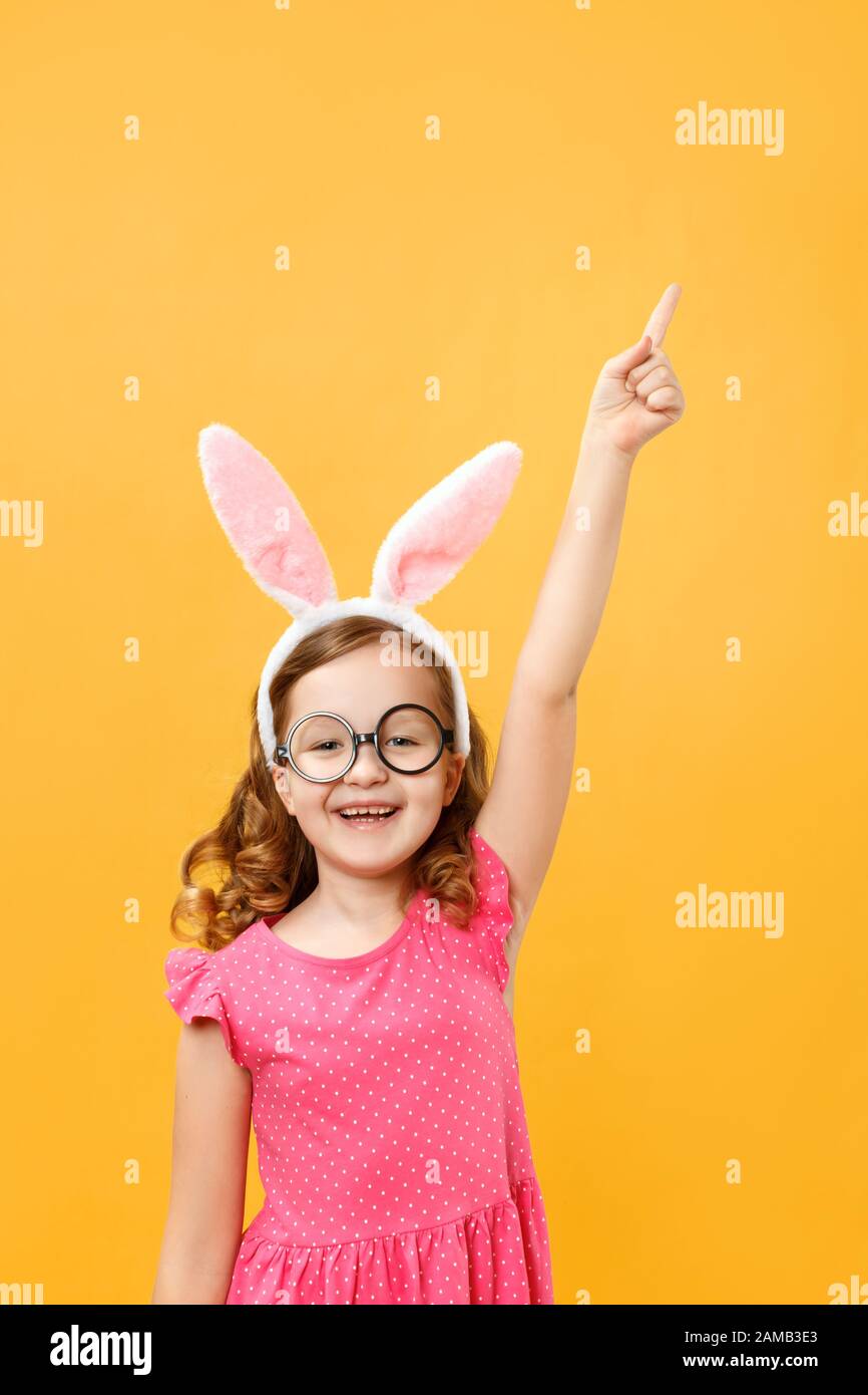 Happy cute little girl in Easter bunny ears and glasses on a yellow background. The child is pointing up at an empty place for text. Stock Photo