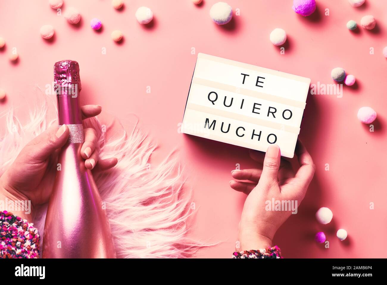 Valentine day creative flat lay, top view. Hands holding lightbox with text and pink bottle of champagne on abstract pink background. Text 'Te quiero Stock Photo
