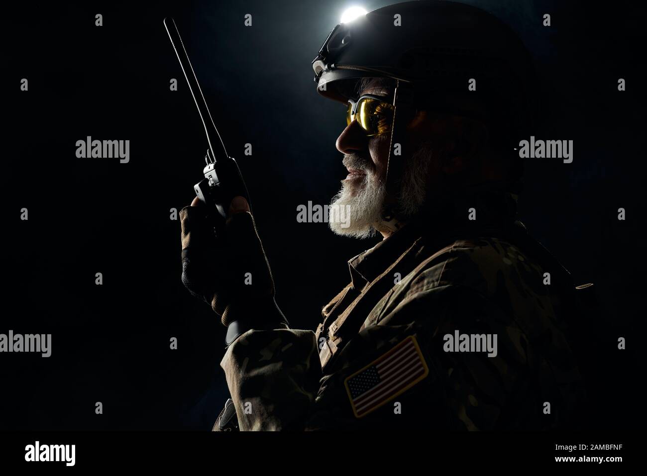 Side view of old military soldier in uniform holding portable radio station in darkness. Close up of male commander in camouflage uniform giving orders. Concept of army, military. Stock Photo