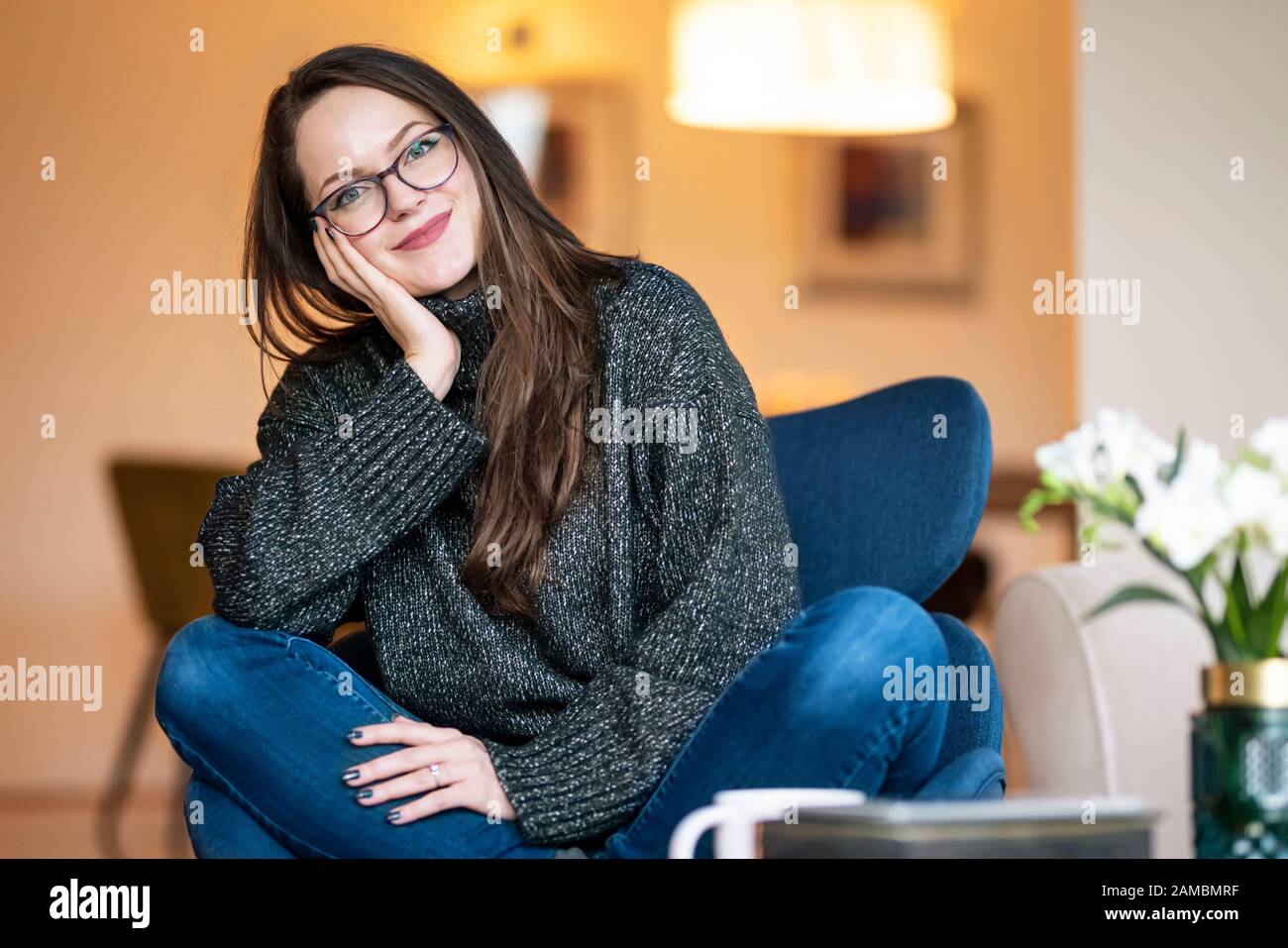 Portrait shot of smiling beautiful brunette young woman relaxing on the chair in the living room at home. Stock Photo