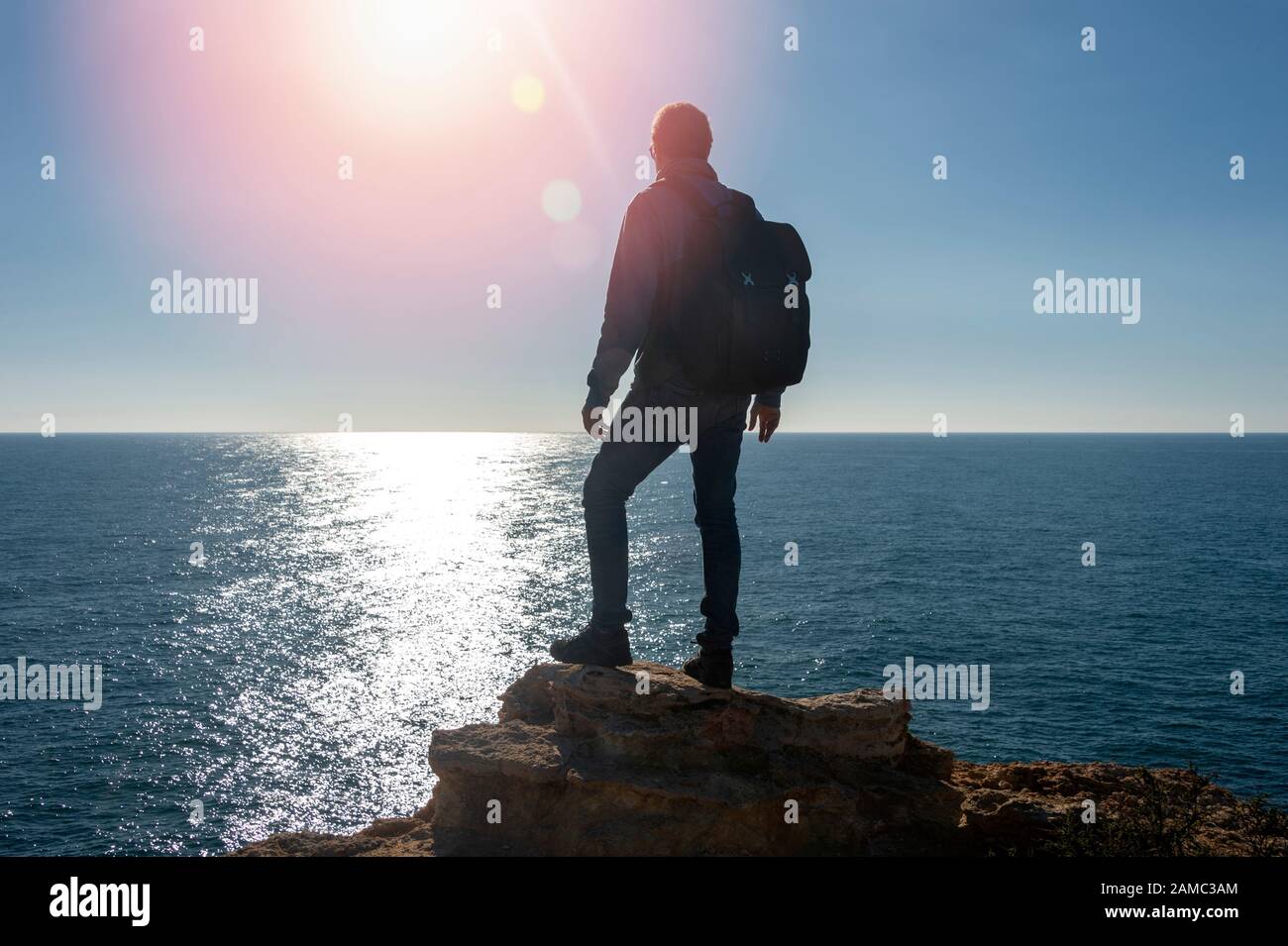 backpacker standing on top of a rock by the sea, lens flare. Stock Photo